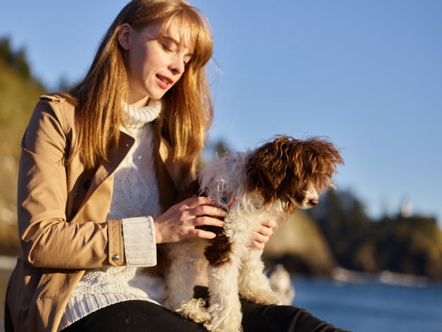 A girl sitting near the ocean holding her white and brown Labradoodle.